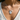 A close-up of a person wearing a delicate silver chain necklace with three charms: a letter 'D', a small silver bead, and an oval medallion pendant, styled with a white V-neck top.